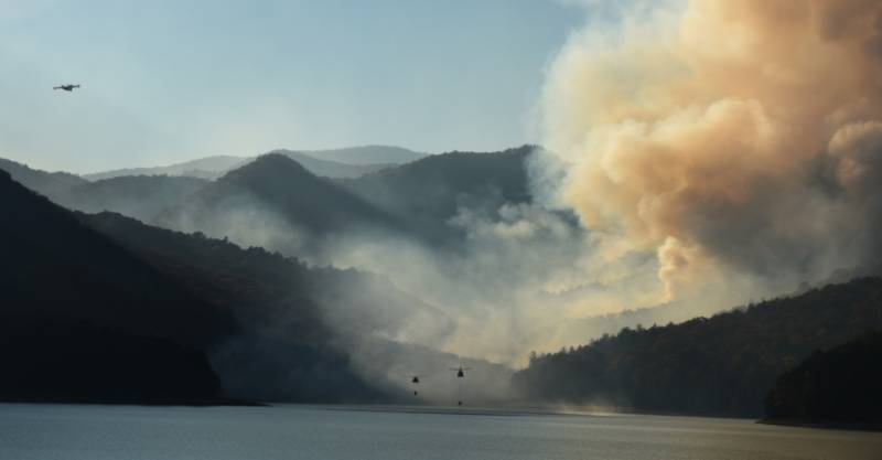 Helicopters scooping water from reservoir with fire in the background.